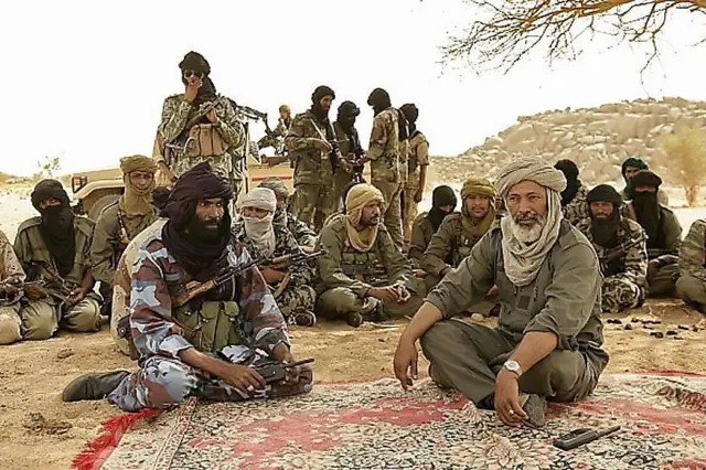 Combattants touaregs discutant sous un arbre, entourés d'armes et de véhicules